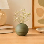 Green vase with white flowers on a wooden surface, with books and a lamp in the background.