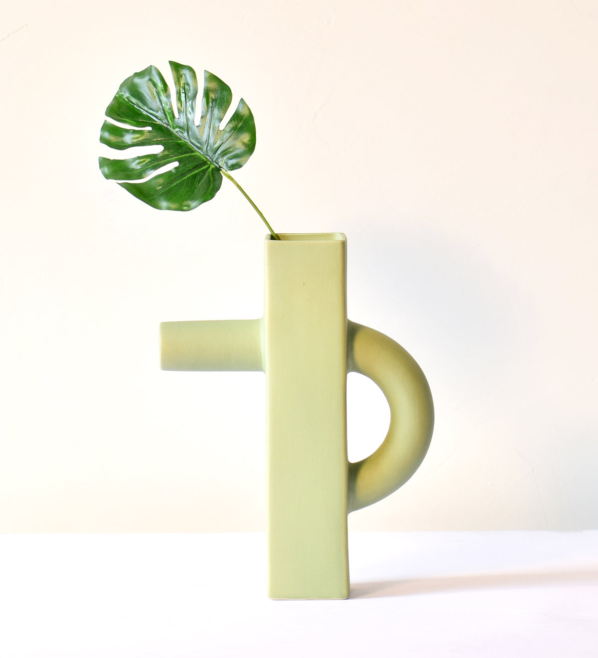 Modern sculptural ceramic flower vase with matte sage green finish styled with tropical leaf in a contemporary luxury interior setting.