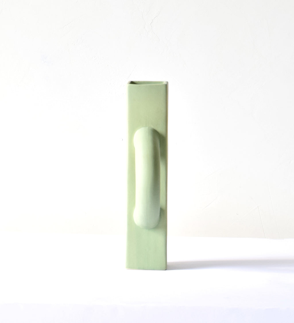 Modern sculptural ceramic flower vase with matte sage green finish styled with tropical leaf in a contemporary luxury interior setting.